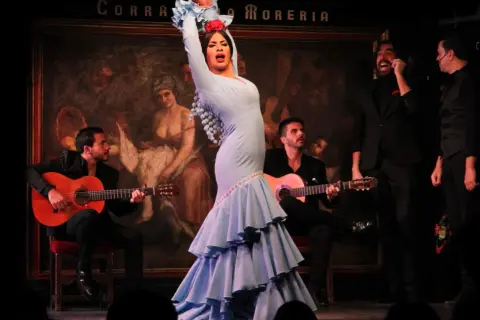 Bailarina de flamenco con vestido azul, guitarristas y cantantes en un escenario. Una escena que evoca el origen de la palabra flamenco.
