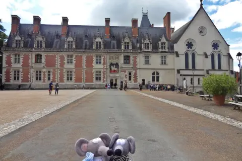 Deux souris en peluche posent devant la majestueuse place du château de Blois, avec son architecture Renaissance et gothique.