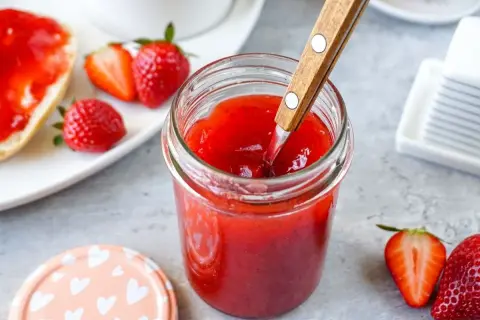 Ein Glas mit leuchtend roter Erdbeermarmelade, die mit einem Holzlöffel umgerührt wird. Daneben frische Erdbeeren und ein halbes Brötchen mit Marmelade – Erdbeermarmelade mit Pfiff für den perfekten Start in den Tag.