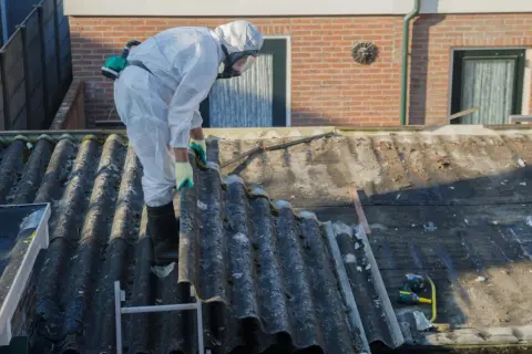 Pracownik w kombinezonie ochronnym usuwa eternit z dachu, dbając o bezpieczeństwo przy pracy z materiałem zawierającym azbest.