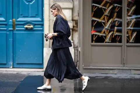 Elegancka kobieta w czarnej, plisowanej spódnicy i marynarce, spacerująca po ulicy. Stylizacje z plisowaną spódnicą są zawsze modne.