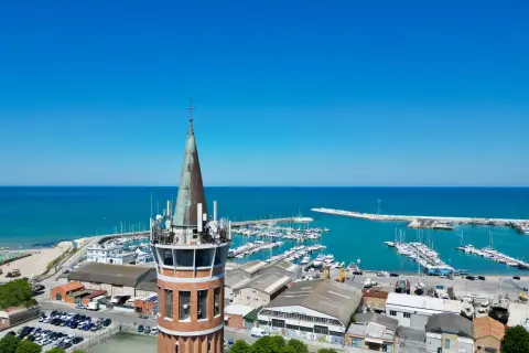 Torre con antenne domina il porto civitanova, pieno di barche e con il mare azzurro sullo sfondo.