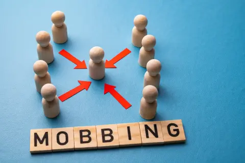 Drewniane figurki ludzi otaczają jedną, a czerwone strzałki wskazują na nią. Słowo "MOBBING" ułożone z liter. Jak udowodnić mobbing w pracy?