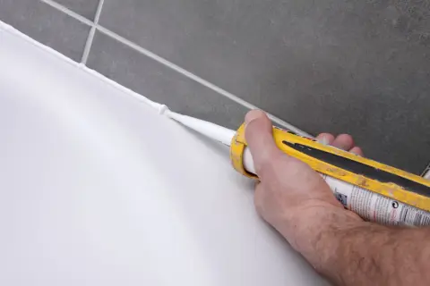 Ręka z pistoletem wyciska biały silikon, wykonując fugowanie płytek.