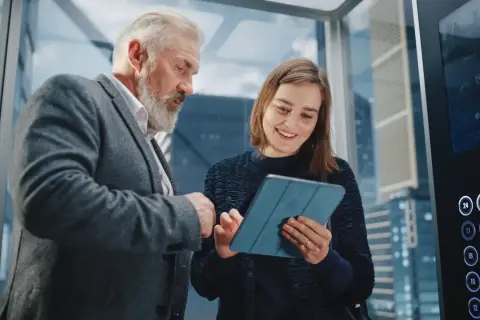 Starszy mężczyzna i młoda kobieta omawiają projekt na tablecie w windzie. To idealny moment na ich elevator pitch.
