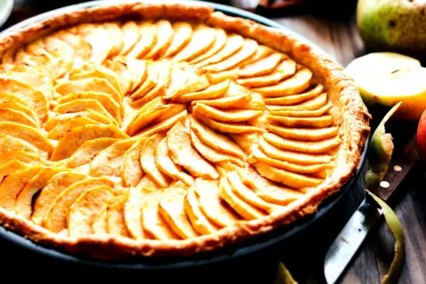 Ein wunderschönes Apfeltarte Rezept, das mit dünn geschnittenen Äpfeln belegt ist. Daneben liegen frische Äpfel und ein Messer.