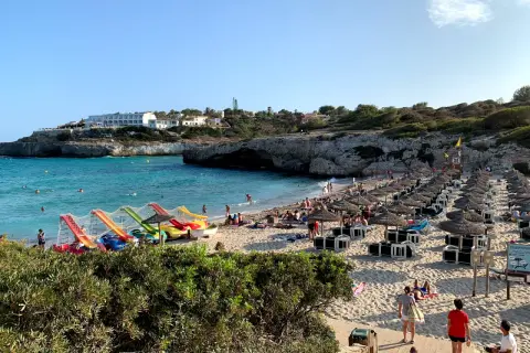 Słoneczna plaża Cala Domingos tętni życiem. Woda jest turkusowa, a na piasku widać rzędy parasoli i leżaków. Dzieci bawią się na kolorowych zjeżdżalniach.