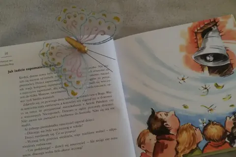 Otwarta książka z ilustracją dzwonu i dzieci patrzących w górę. Motylowa zakładka ozdabia stronę z tekstem o tym, jak ludzie zapomnieli o Bogu.