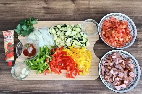 Pokrojone warzywa (papryka, cukinia, cebula, pomidory), kiełbasa, czosnek, przyprawy i koncentrat pomidorowy na desce.