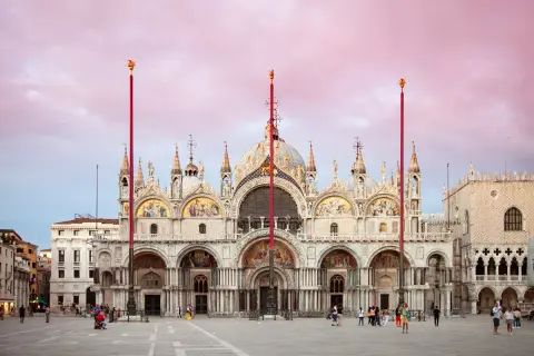 La maestosa Basilica di San Marco a Venezia, con le sue cupole dorate e mosaici scintillanti, si staglia contro un cielo rosa al tramonto.
