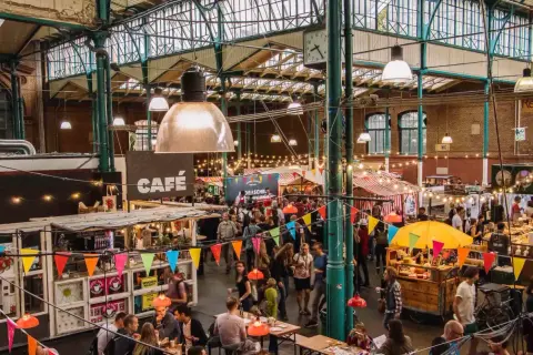 Buntes Treiben auf einem Street Food Festival in Berlin. Menschen schlendern zwischen Ständen mit Leckereien unter einer beeindruckenden Glasdachkonstruktion.