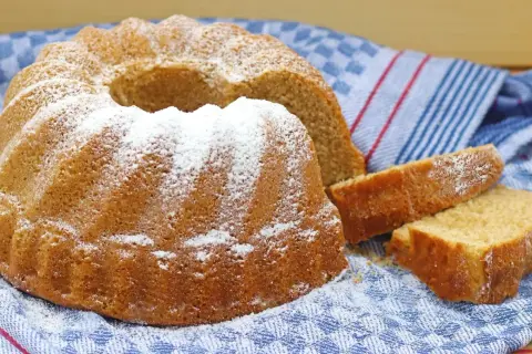 Puszysta babka z owocami, oprószona cukrem pudrem, czeka na spróbowanie. Obok leżą dwa kawałki ciasta.