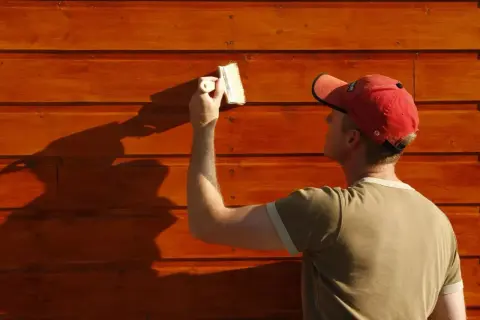 Un homme en casquette rouge est en train de peindre du bois brut extérieur avec un pinceau. La lumière du soleil projette une ombre nette sur le mur.