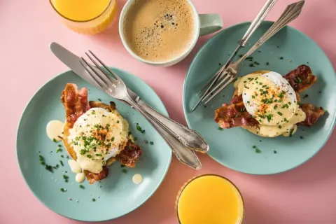 Zwei Teller mit **Egg Benedict**, garniert mit Speck und Schnittlauch, daneben Kaffee und Orangensaft.