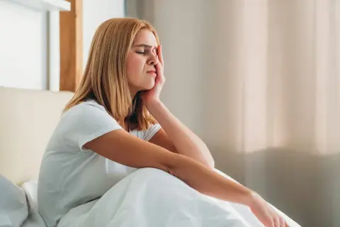 A young woman woke up with a headache, grimacing and holding her forehead as she sits in bed.