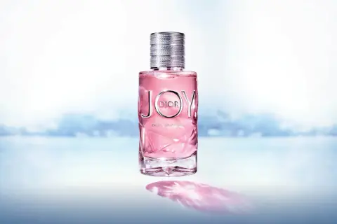 Różowy flakon Dior Joy 90ml z metalową nakrętką, na tle rozmytego, błękitnego krajobrazu.