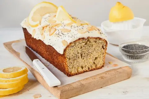 Ein saftiger Zitronen Mohn Kuchen mit weißem Zuckerguss, Mandelsplittern und Zitronenscheiben. Daneben liegen frische Zitronenscheiben und Mohn.