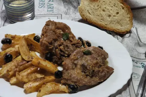 Un saporito ossobuco di tacchino con patate arrosto e olive nere, accompagnato da pane fresco e un bicchiere di vino bianco.
