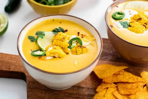 Zwei Schüsseln mit cremiger Mais-Suppe, garniert mit Maiskolben, Limette und Jalapeños, daneben Tortilla-Chips.