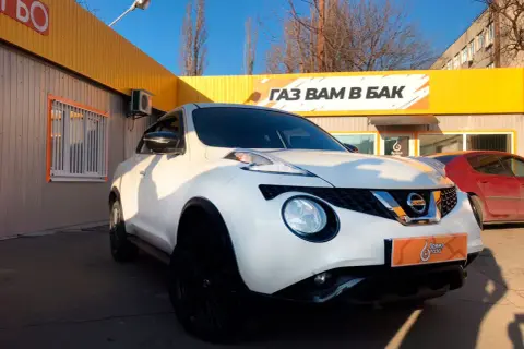 Biały Nissan Juke 1.6 benzyna z czarnymi felgami przed budynkiem z żółtym banerem "GAZ WAM W BAK".