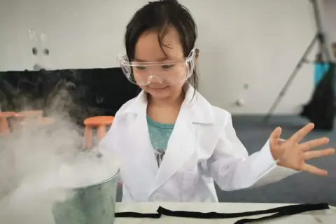 Niña con bata y gafas realiza un experimento científico para niños, creando humo blanco en un cubo metálico.