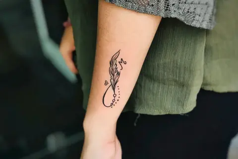 Ein kleines Feder-Tattoo mit Unendlichkeitszeichen und dem Buchstaben M auf dem Unterarm eines Mannes.