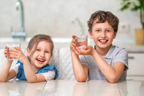 Uśmiechnięta dziewczynka i chłopiec trzymają szklanki z wodą. Cieszą się orzeźwiającym smakiem, jakby pili miękką wodę prosto ze źródła.