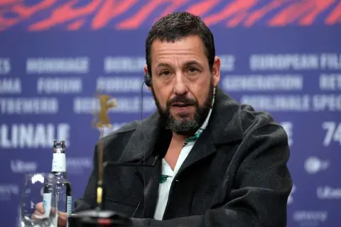 Adam Sandler, känd från många roliga filmer, sitter vid ett bord med en mikrofon och en vattenflaska.