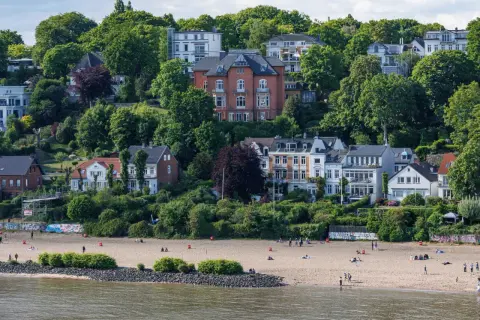 Prächtige Villen und grüne Hügel über einem Sandstrand an den Elbvororten Hamburgs. Menschen genießen den sonnigen Tag am Wasser.