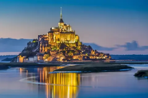 Mont Saint-Michel lyser upp i skymningen, en dröm för en semester i Frankrike. Vattnet speglar dess magiska ljus.