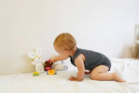 A baby explores Montessori baby activities, reaching for a teddy bear among soft toys on a white blanket.