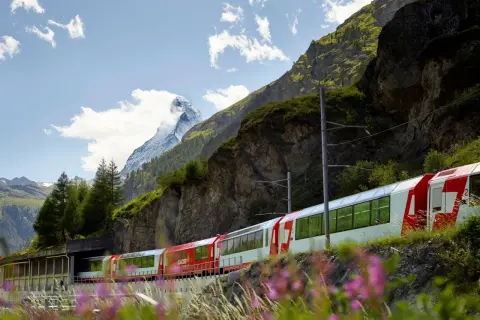 Czerwono-biały pociąg Glacier Express mija skaliste zbocze górskie z ośnieżonym szczytem w tle.
