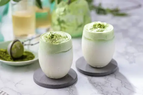 Deux verres de dalgona matcha, une boisson crémeuse et verte, sur des sous-verres gris.