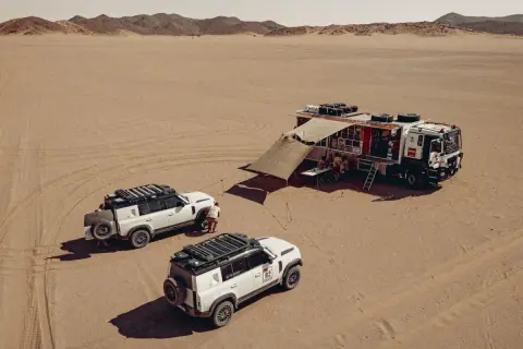 Un camion Dakar et deux 4x4 blancs dans le désert. L'équipe se réunit sous un auvent, prête pour l'aventure.