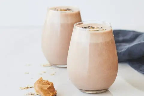 Två glas med krämig choklad- och jordnötssmoothie, toppad med chiafrön. En sked med jordnötssmör och havregryn ligger bredvid. En perfekt start på dagen, som Don Pedro skulle ha gillat.