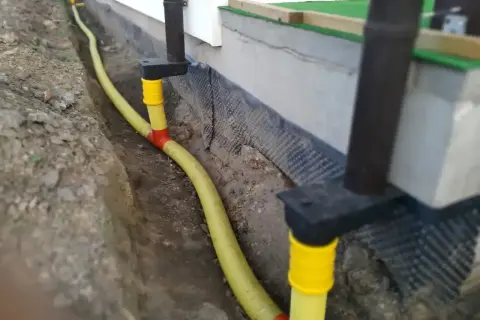 System odprowadzenia wody z rynny, z żółtą rurą biegnącą w wykopie przy fundamencie.