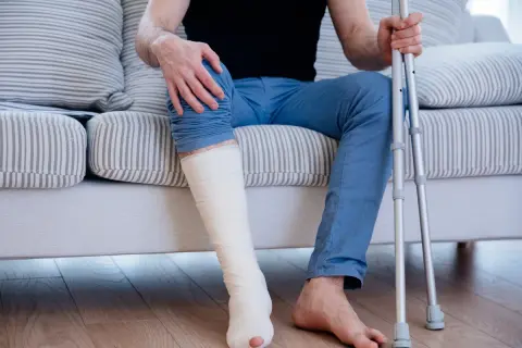 Mężczyzna siedzi na kanapie z zabandażowaną, złamaną nogą i kulami.