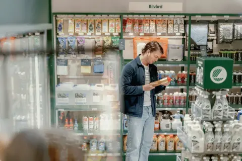 Man i blå jacka och jeans tittar på en produkt i en butik med bilvårdsprodukter. Kanske undrar han vad som menas med "sae olja" för att välja rätt.