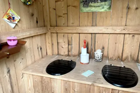Två svarta toalettlock på en träbänk i ett utedass. En rulle toalettpapper och en lykta står på bänken. En bok med "utedass regler" hänger på väggen.