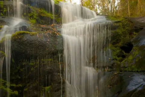 Cascada en los cañones del río Lozoya, con rocas cubiertas de musgo y hojas secas. El agua cae en velos blancos sobre la piedra oscura.