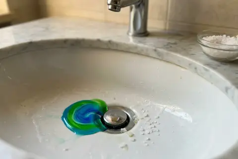 Un lavandino con un vortice di sapone blu e verde vicino allo scarico, e del sale grosso per sturare lavandini sparso sul bordo.