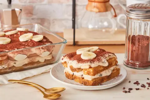 Ein köstliches Bananen Tiramisu, geschichtet mit Creme und Bananenscheiben, bestäubt mit Kakao. Daneben ein Glas Kakaopulver.