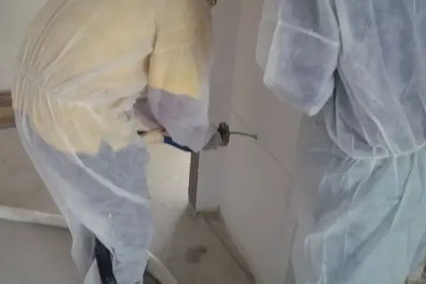 Dos operarios con trajes protectores aplican espuma intumescente en una pared, sellando una junta para protección contra incendios.