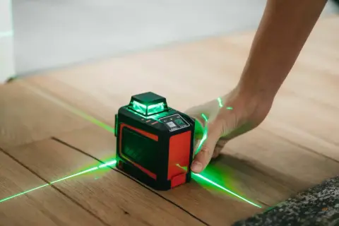 Poziomica laserowa z zielonymi liniami na drewnianej podłodze, obsługiwana przez dłoń.