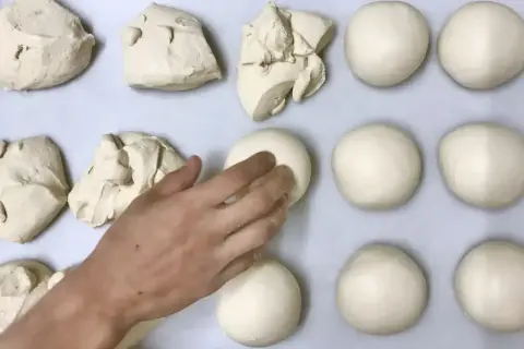 Des boules de pâte à pizza sont disposées sur une plaque. Une main façonne délicatement l'une d'elles.