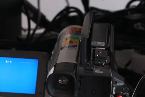 En videokamera med öppen lucka och en blå skärm. Kablar syns i bakgrunden, en påminnelse om analoga bilder och äldre teknik.