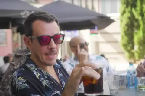 Un hombre con gafas de sol rojas y camisa tropical bebe una bebida en una terraza, quizás disfrutando de un momento de pantomima full Zaragoza.
