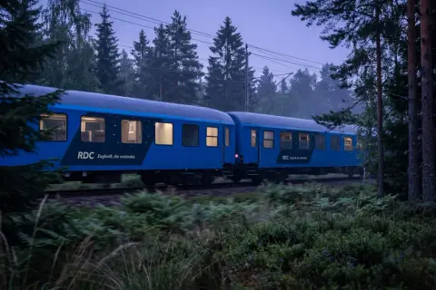 Blått nattåg från RDC passerar genom en skog i skymningen. Tåget är på väg mot Berlin, en resa som börjar i Sverige.