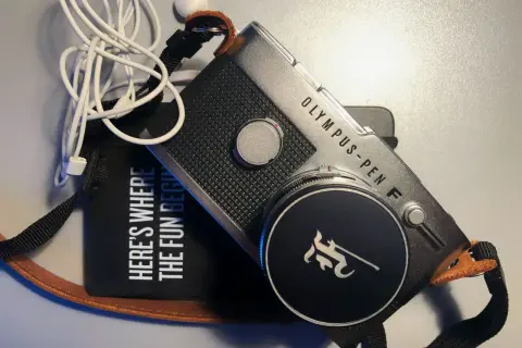 The Olympus Pen FV camera, with its iconic design, sits beside a black pouch that reads "HERE'S WHERE THE FUN BEGINS." White earbuds are tangled nearby.