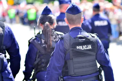 Policjanci w mundurach, tworzący zgraną kartę rodziny mundurowej, patrolują ulicę. Na kamizelkach taktycznych widnieje napis "POLICJA".